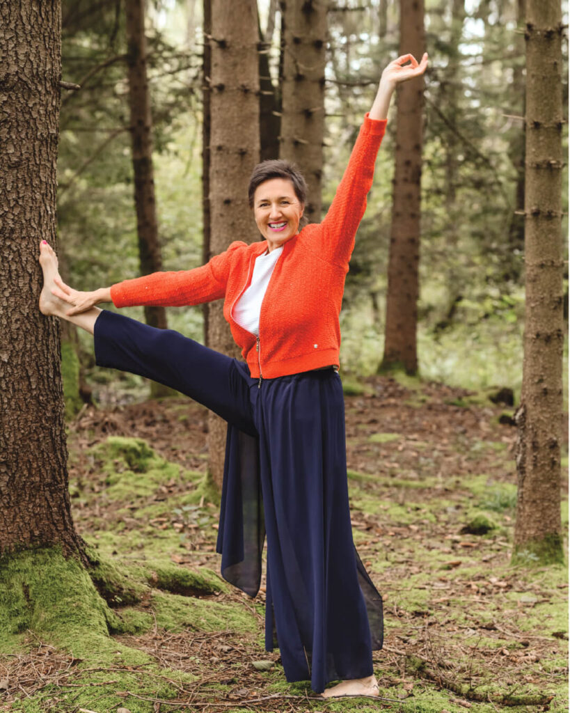 Karin-Buttinger-Selbstliebe-Kurs-Venus-erwacht-Karin-steht-lachend-im-Wald-ein-Bein-auf-Kopfhoehe-an-einen-Baum-gelehnt